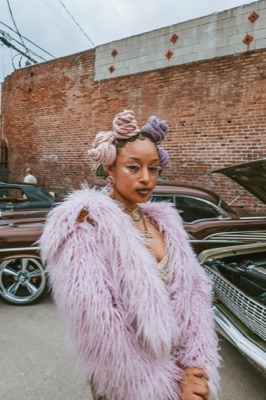 Model in a pink fur coat and pastel bantu knots at a lowrider car show
